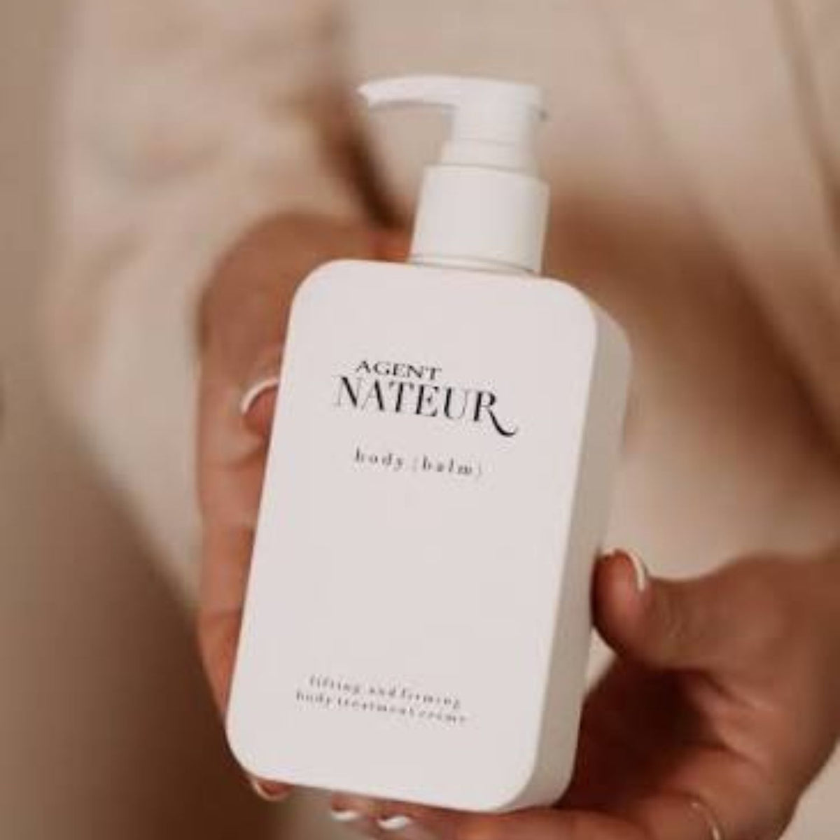 Hands holding a white pump bottle of Agent Nateur Body (Balm) body treatment crème.