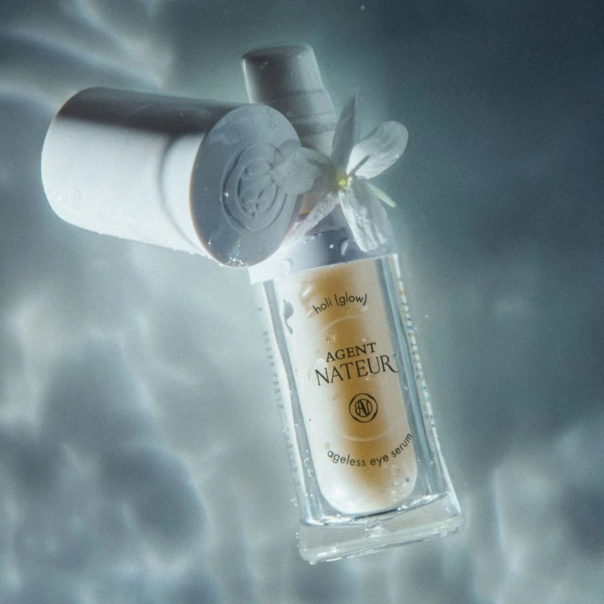 Agent Nateur Holi (Glow) Ageless Eye Serum bottle with cap removed beside a white flower in rippled light.