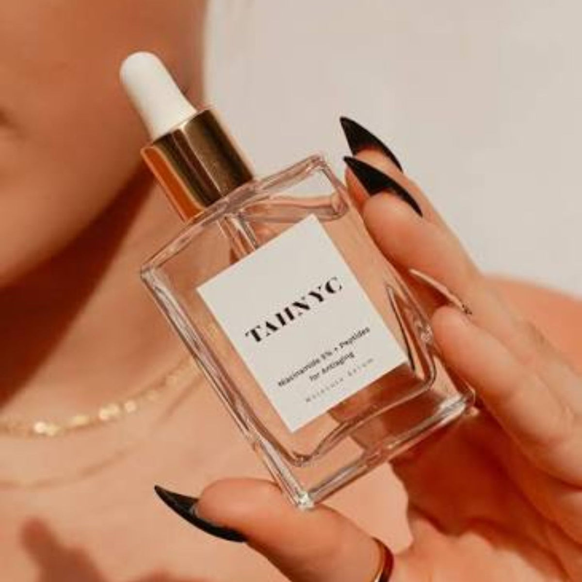 Hand holding a clear glass dropper bottle of TAHNYC Niacinamide 5% + Peptides serum.