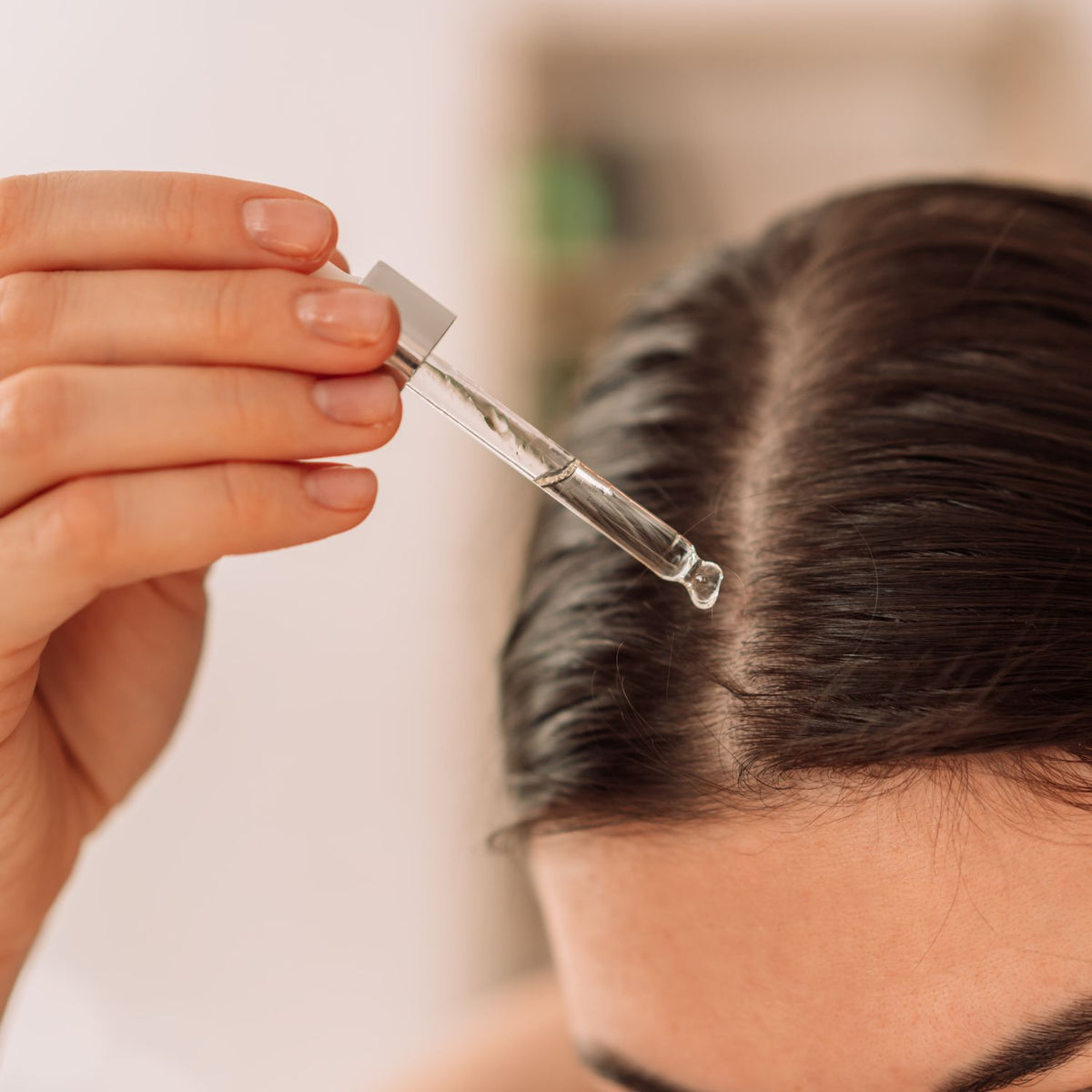Hair serum being applied with a glass dropper along a parted scalp.